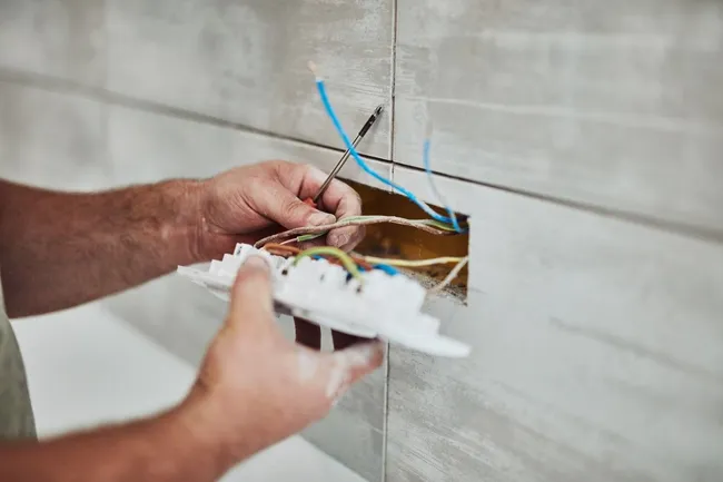 Hands wiring an electrical box in a tiled wall, with blue and white wires exposed.
